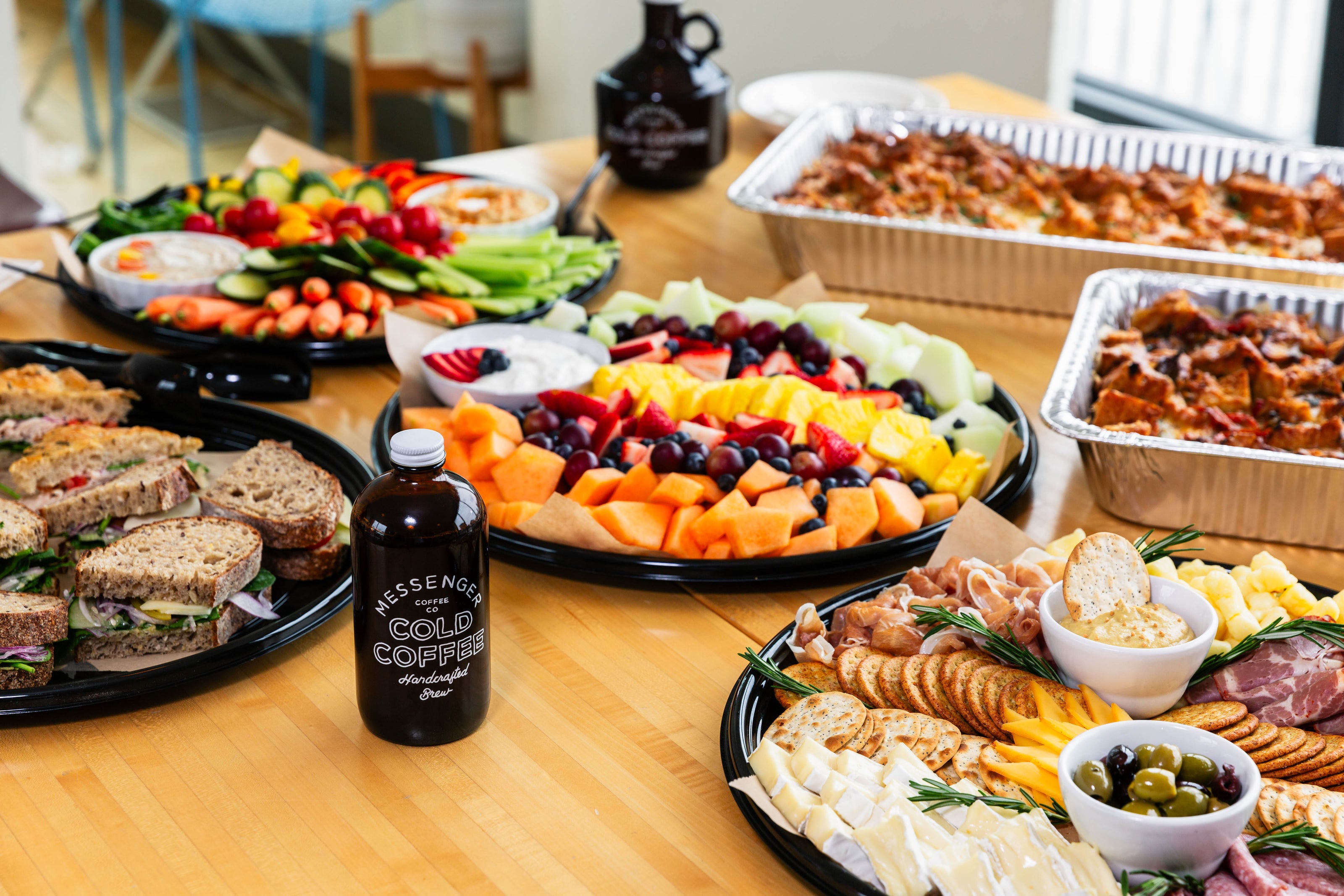 Catering spread with sandwiches, fruit platter, cheese and crackers, vegetables, and trays of pasta on a wooden table